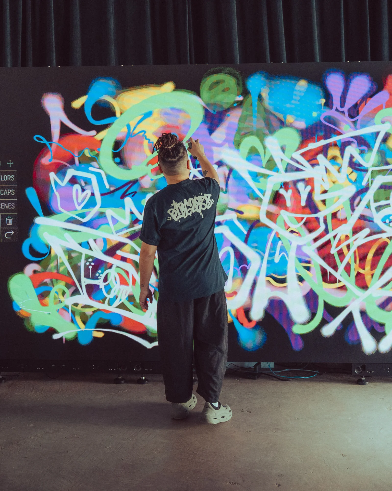 Writer in a 'BURNOUT' tee painting on the Graffiti+ wall at MGM Cotai with the menu UI (COLORS, CAPS, STICKERS, SCENES) visible on the left
