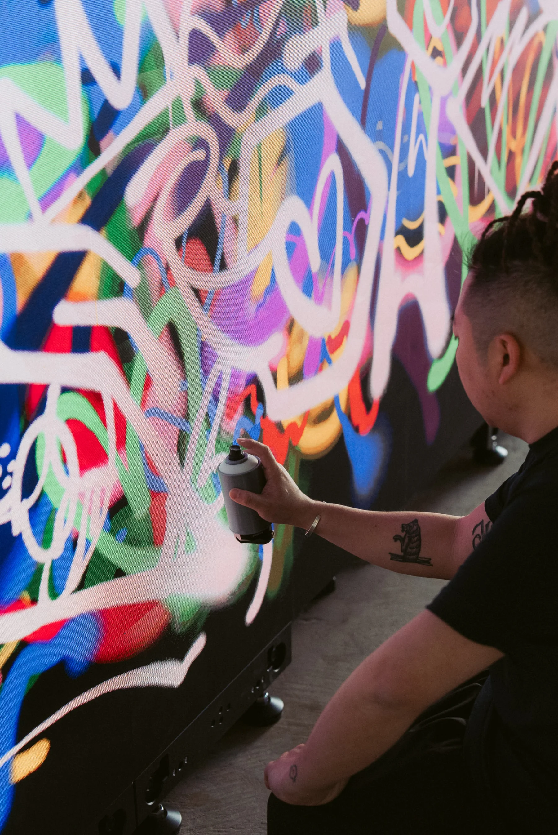 Close-up of a Macau graffiti writer spraying white throw-up tags across the Graffiti+ wall at MGM Cotai — visible tattoos on forearm