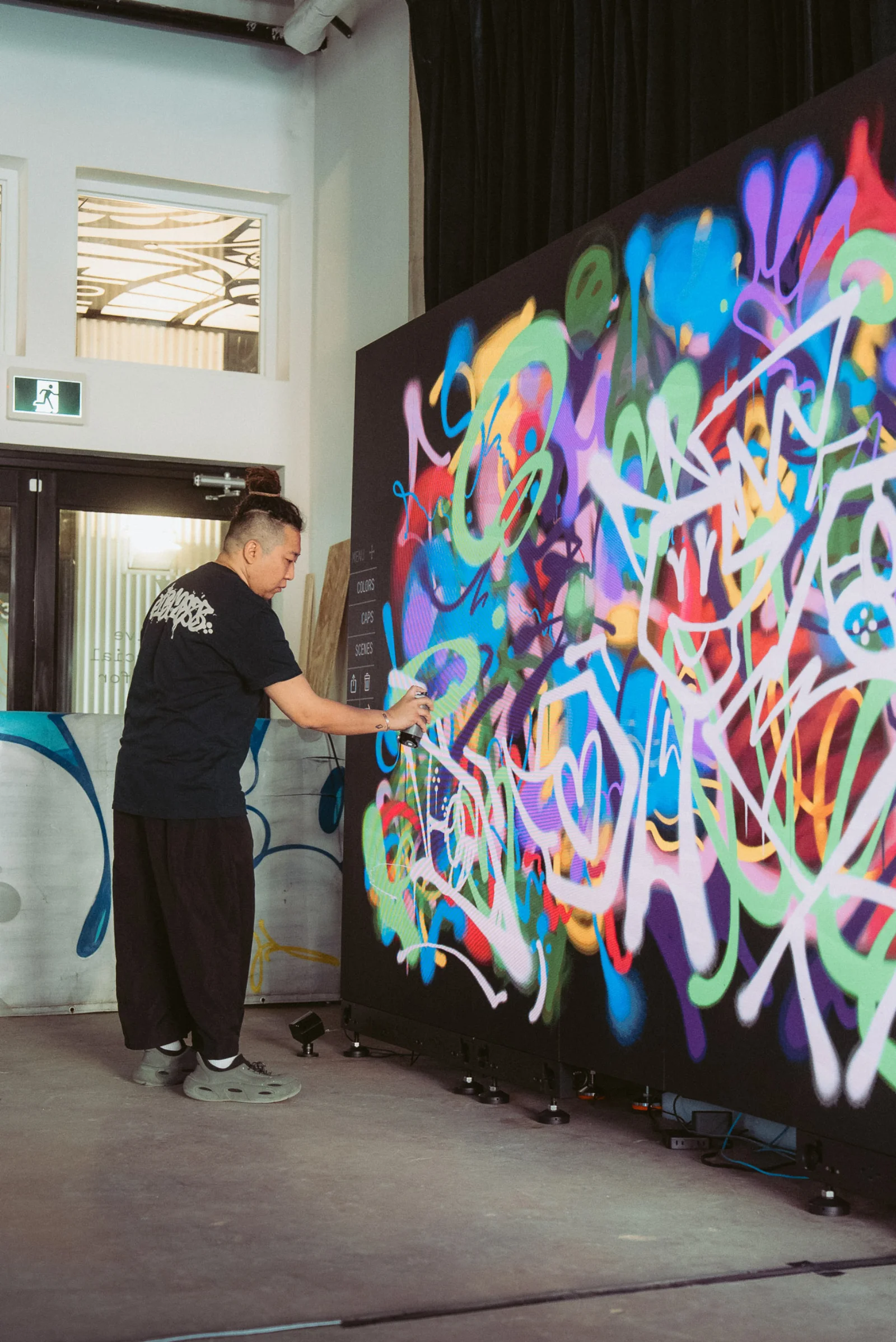 Wide shot of a writer painting on the Graffiti+ wall at MGM Cotai — finished piece covered in white throw-up tags over a colourful background