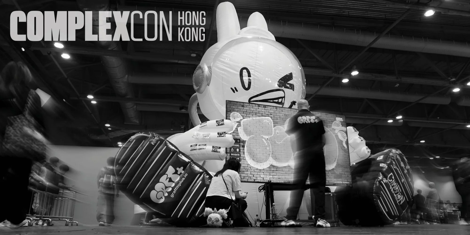 The 'Digital Graffiti Space Bun' installation at ComplexCon Hong Kong 2025 — wide black-and-white view of the 13-foot Billion Buns inflatable Space Bun embracing a 100-inch 4K Graffiti+ × Chairman Ting digital graffiti canvas, with 'COMPLEXCON HONG KONG' logo overlay