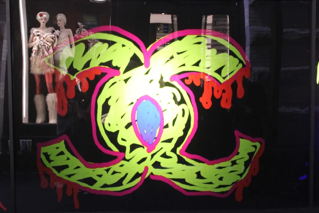 The interlocking Chanel double-C logo rendered in neon green, pink and blue graffiti on the interactive LED wall at the Chanel SoHo flagship, with Chanel mannequins visible behind, 2010