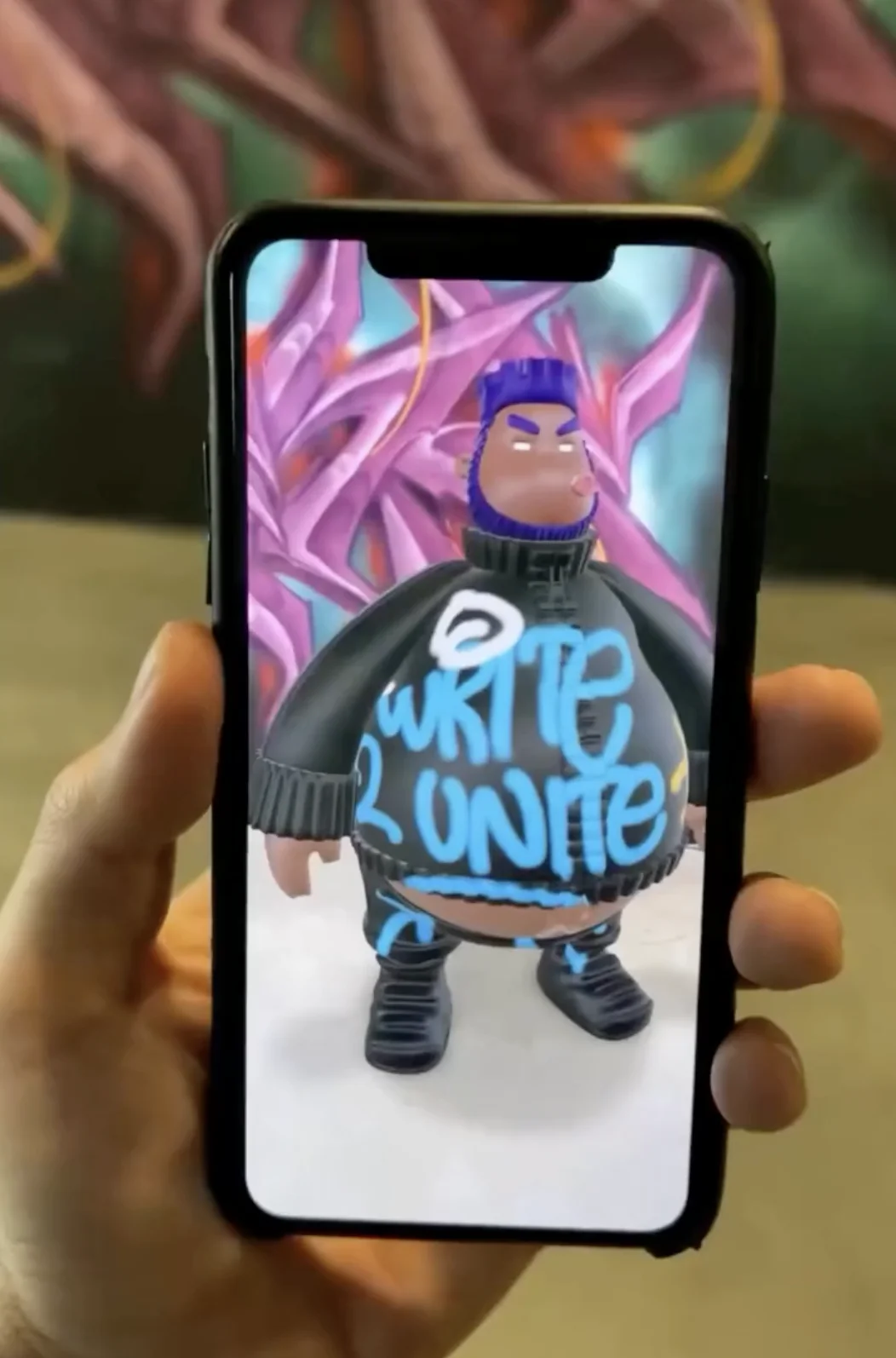 Phone showing the painted 'WRITE 2 UNITE' designer toy as an AR character on the DesignerCon 2024 show floor — Graffiti+ QR-to-AR take-home