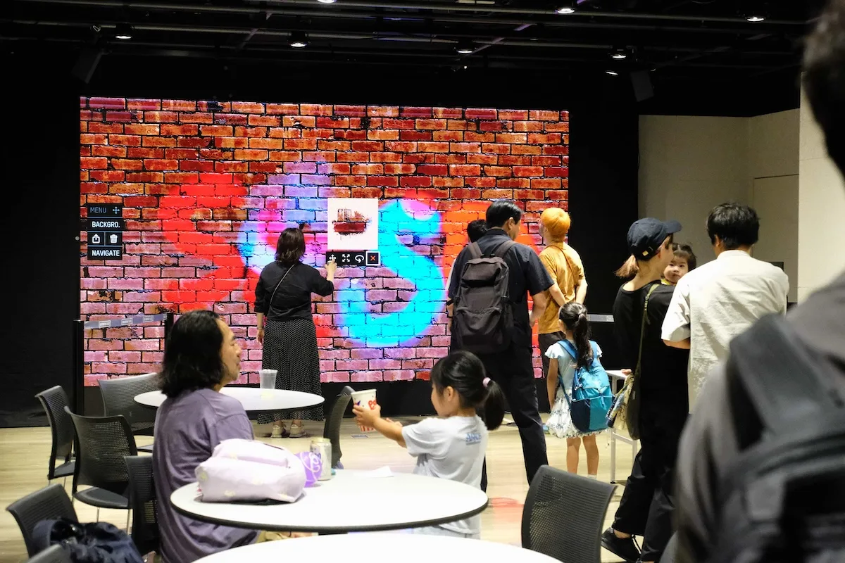 The Graffiti+ digital spray art LED wall at BON CAST., Shibuya Cast — a guest painting on the brick-textured screen while festival-goers and families watch