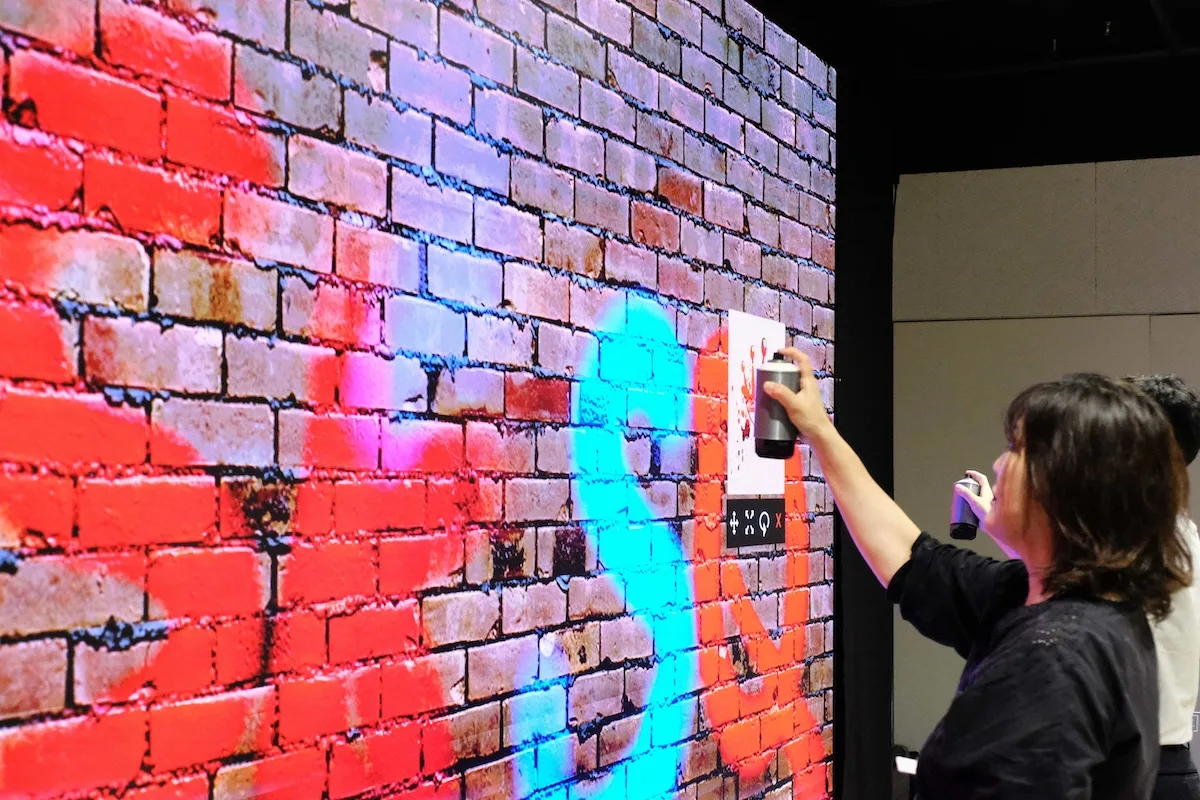 Close-up of a BON CAST. guest holding a physical spray can and painting a piece on the Graffiti+ LED wall, with the on-screen colour palette and tools visible