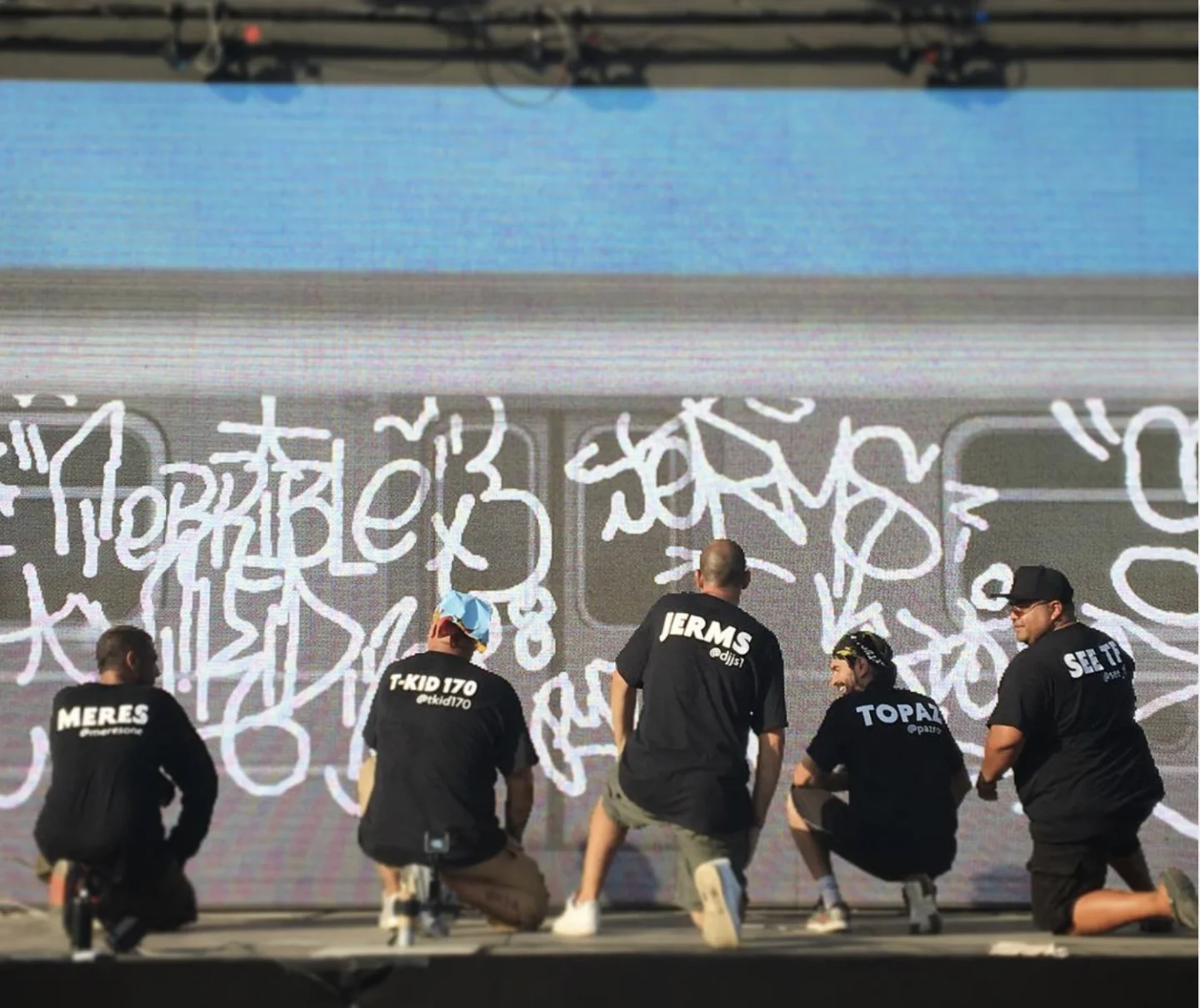 OG New York writers Meres One, T-Kid 170, Jerms, Topaz and See kneeling on stage, tagging a full-size subway car on the Graffiti+ LED wall at Panorama Festival 2016
