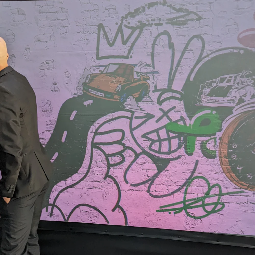 Carson Ting (Chairman Ting) studying his collaborative digital mural on the Graffiti+ wall at Porsche Centre Richmond — featuring his signature character work alongside Porsche silhouettes