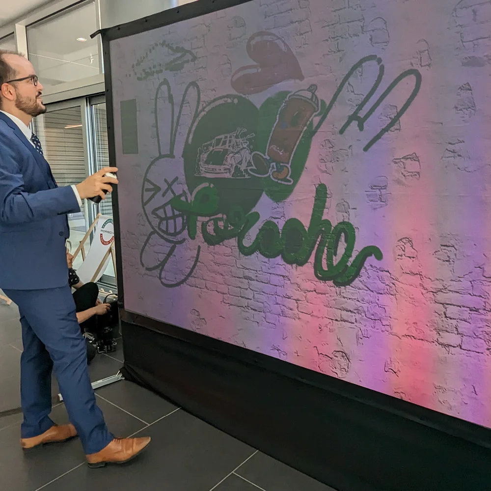 Suited VIP attendee painting 'Porsche' lettering with a custom illustration set on the Graffiti+ LED wall at the Festival of Dreams